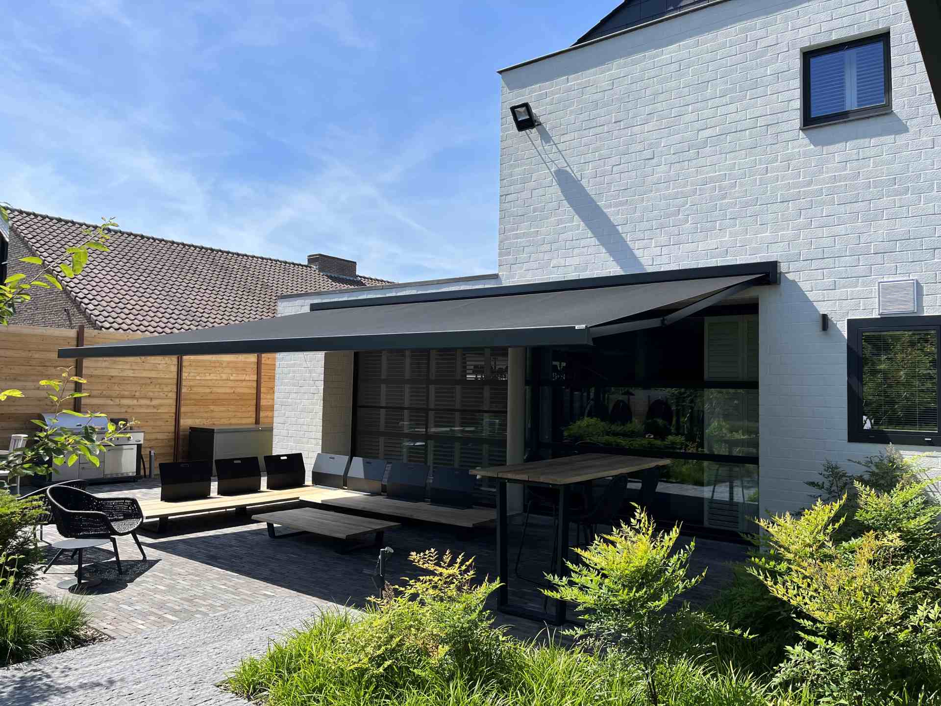 Modern wit bakstenen huis met houten terras, donkergrijs zonnetent en groene planten.